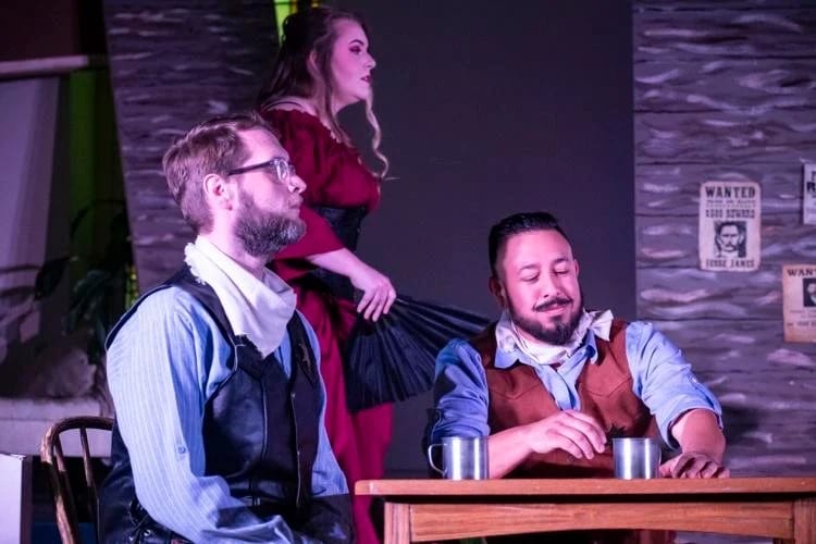 Two actors seated at a saloon table while another performer stands behind them