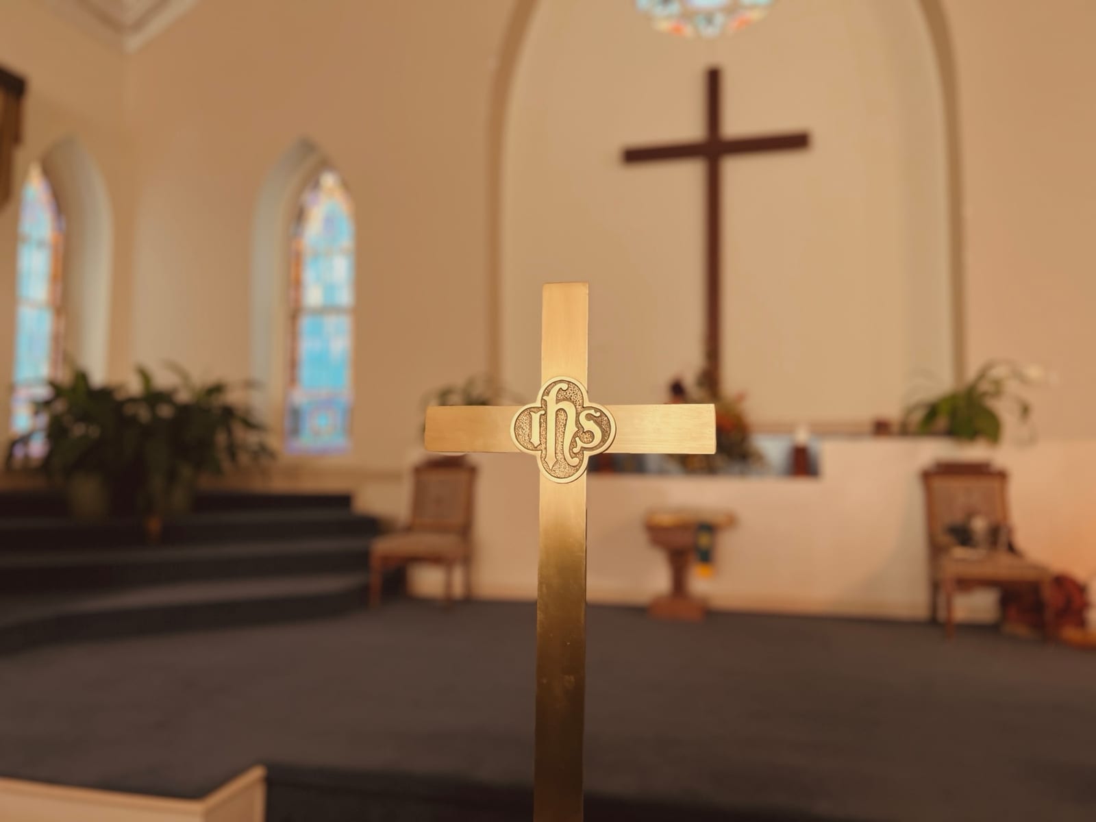 A brass processional cross with the sanctuary cross and stained glass behind it